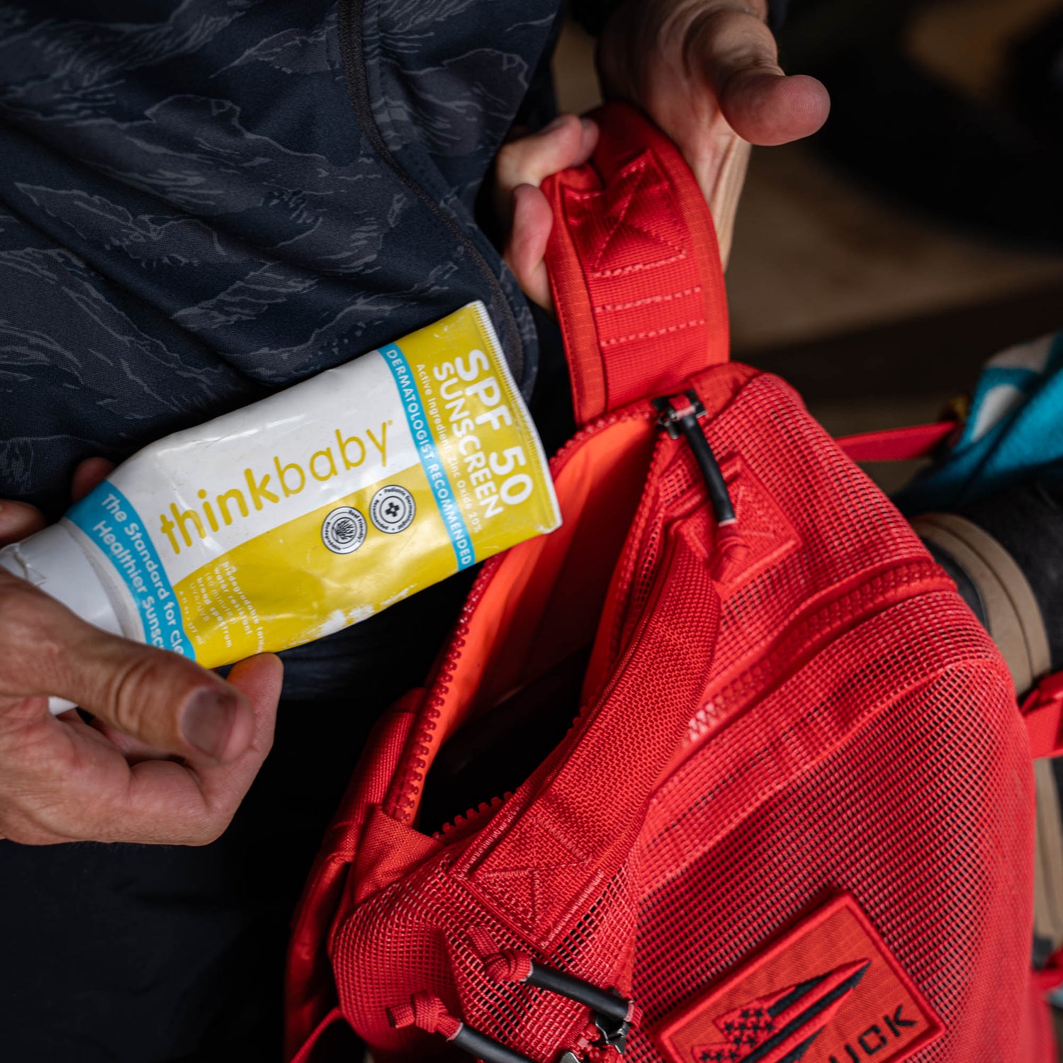 A person is putting thinkbaby SPF 50 sunscreen into a Mesh Ruck red beach backpack, while wearing a dark jacket.