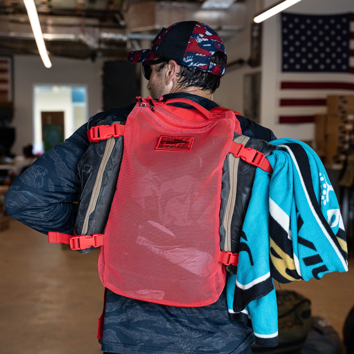 A man with a Mesh Ruck in red and a colorful towel over his shoulder stands indoors near an American flag.
