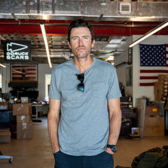 A man wearing the Short Sleeve Pocket Tee - Tri-BlendX stands in a workshop with an American flag and GORUCK SCARS sign in the background, embodying classic simplicity and a versatile spirit.