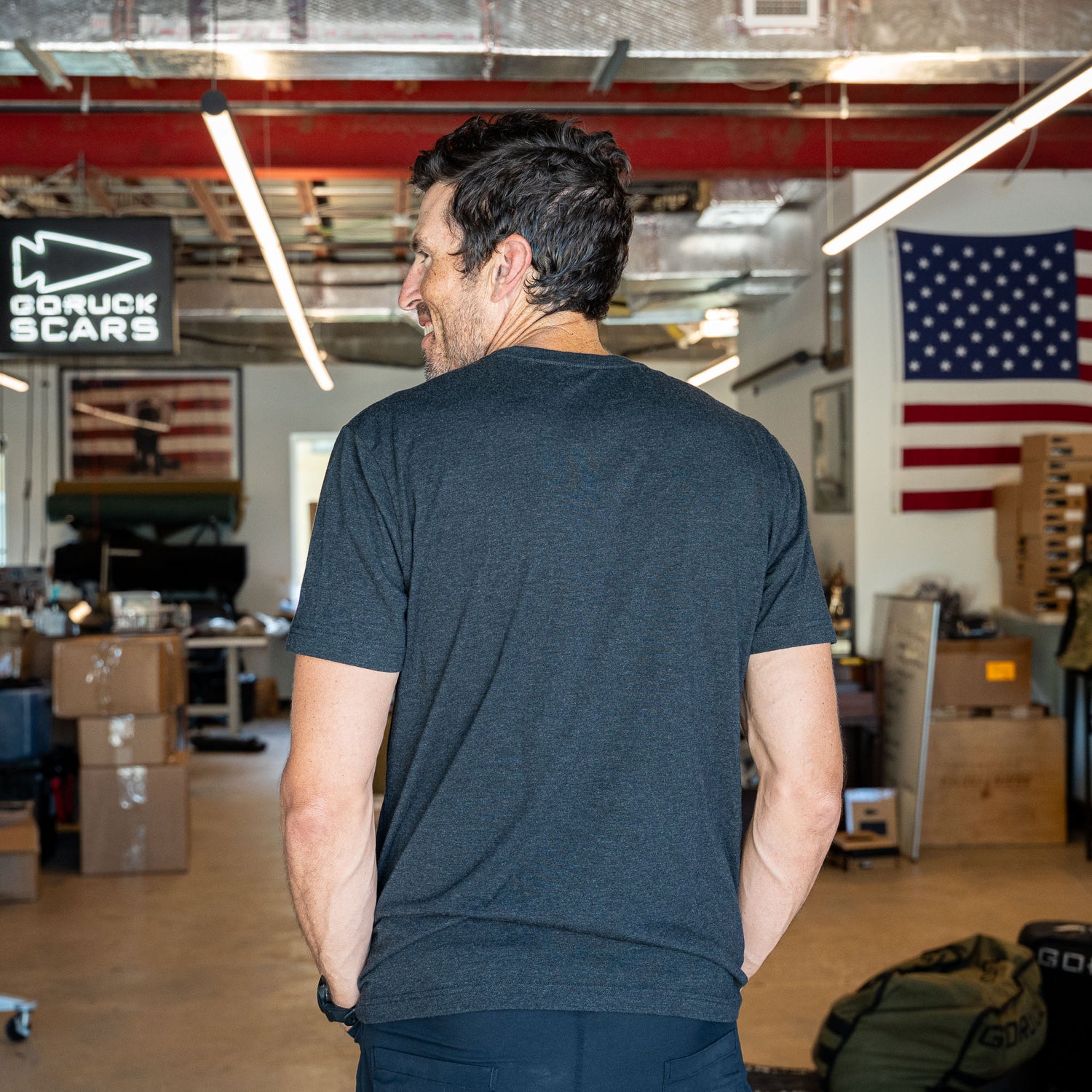 A man wearing the Short Sleeve Pocket Tee - Tri-BlendX stands with his back to the camera in an industrial workspace adorned with U.S. flags, embodying classic simplicity and a versatile style that suits any setting.