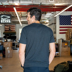 A man wearing the Short Sleeve Pocket Tee - Tri-BlendX stands with his back to the camera in an industrial workspace adorned with U.S. flags, embodying classic simplicity and a versatile style that suits any setting.