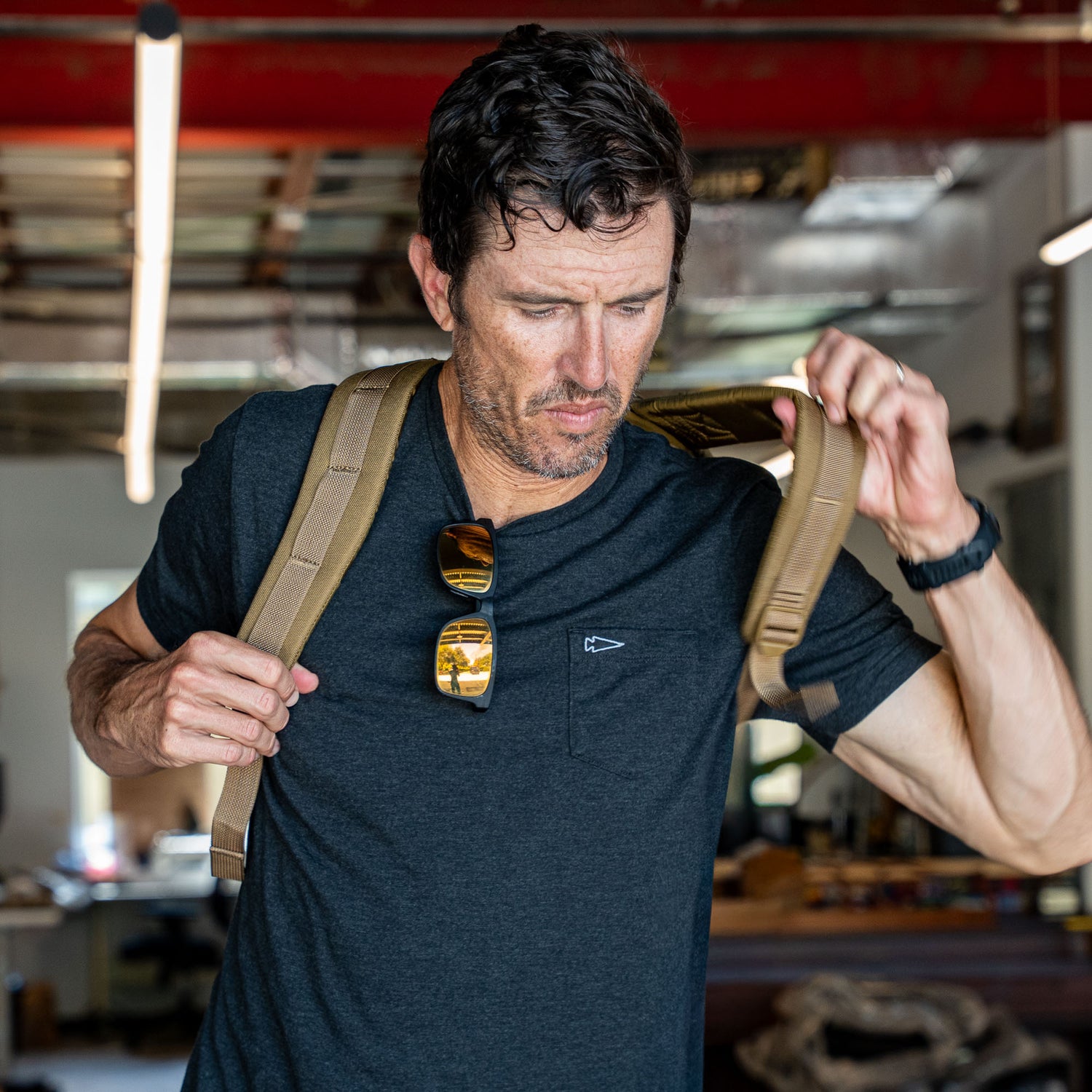 A man in a black shirt puts on the Double Bullet Ruck w/ Bombproof Laptop Compartment - 18L, a tan travel backpack, in an office, with sunglasses hanging from his collar.