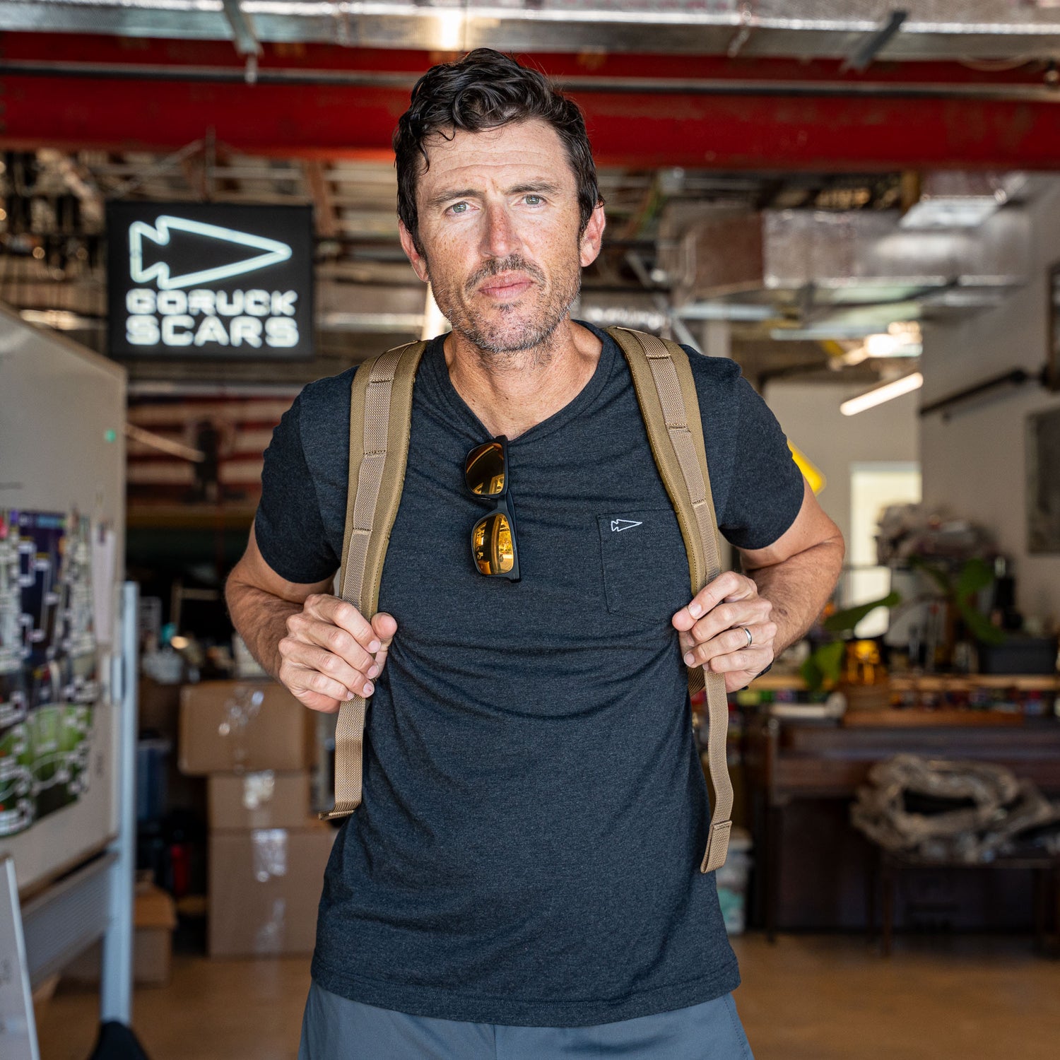 A man wearing a travel backpack and sunglasses on his shirt stands in a workshop with a 