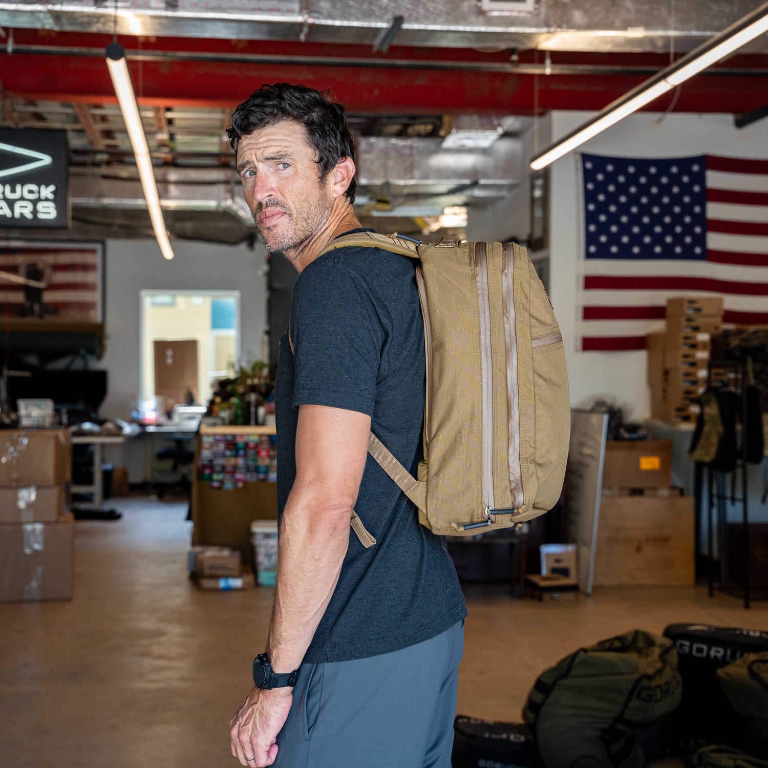 A man stands in an industrial room with boxes and an American flag on the wall, wearing the Double Bullet Ruck w/ Bombproof Laptop Compartment - 18L in brown.