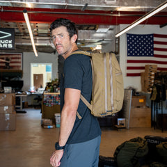 A man stands in an industrial room with boxes and an American flag on the wall, wearing the Double Bullet Ruck w/ Bombproof Laptop Compartment - 18L in brown.