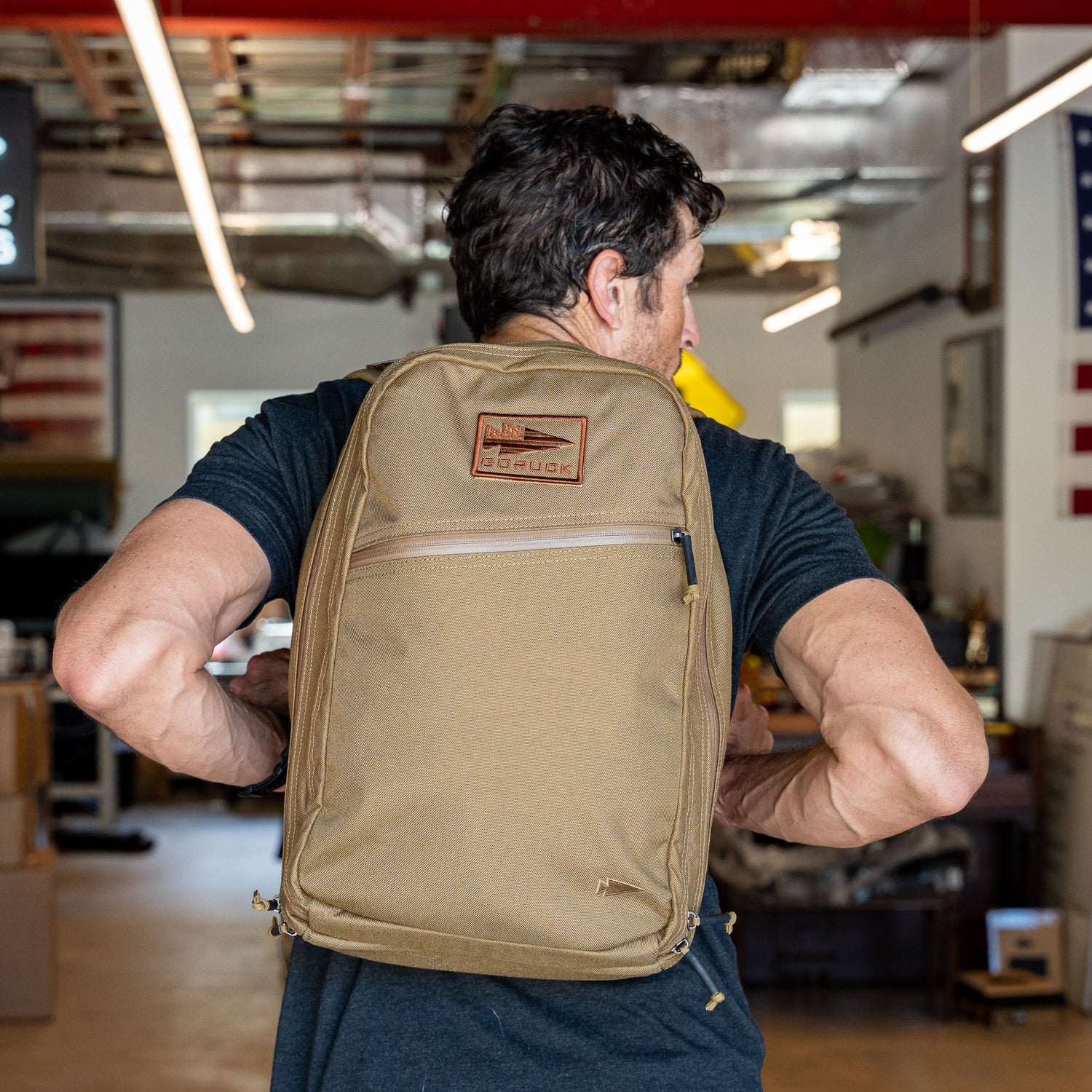 A man wearing a tan Double Bullet Ruck w/ Bombproof Laptop Compartment - 18L travel backpack indoors, facing away, with an American flag visible in the background.