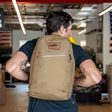 A man wearing a tan Double Bullet Ruck w/ Bombproof Laptop Compartment - 18L travel backpack indoors, facing away, with an American flag visible in the background.