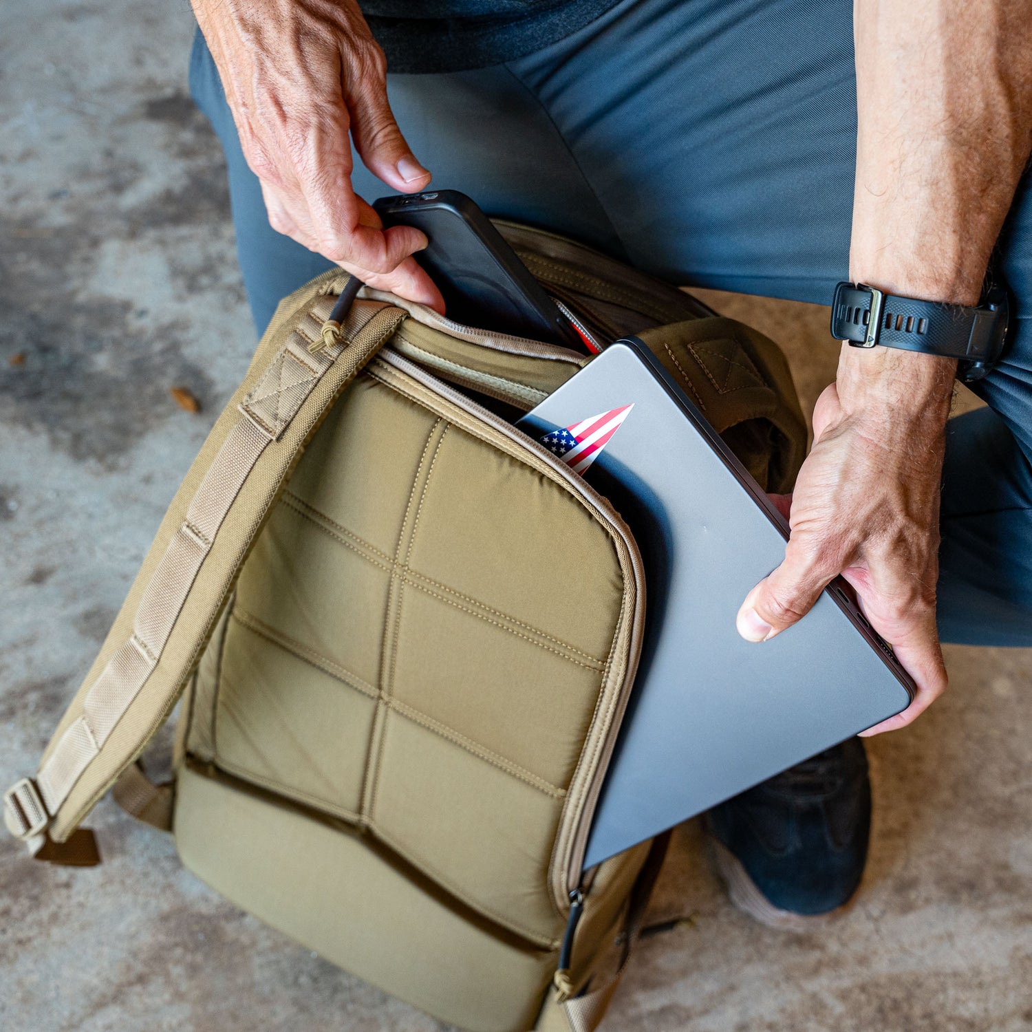 A person kneels on the ground, placing a laptop featuring a US flag sticker into the bombproof laptop compartment of the Double Bullet Ruck w/ Bombproof Laptop Compartment - 18L in tan.