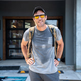 A man wearing sunglasses, a backpack, and the Short Sleeve Pocket Tee - Tri-BlendX in gray stands outside a building, looking at the camera with an air of classic simplicity.