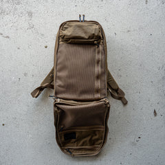 A brown Double Bullet Ruck w/ Bombproof Laptop Compartment - 18L lays open on a concrete surface, revealing its mesh inner compartments, durable zippers, and bombproof laptop compartment built for secure travel.