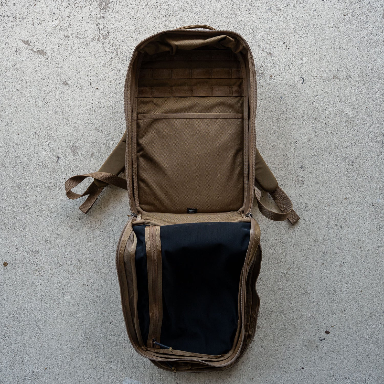 An empty brown Double Bullet Ruck w/ Bombproof Laptop Compartment - 18L travel backpack lies open on a concrete surface, displaying its bombproof laptop compartment along with its interior compartments and straps.