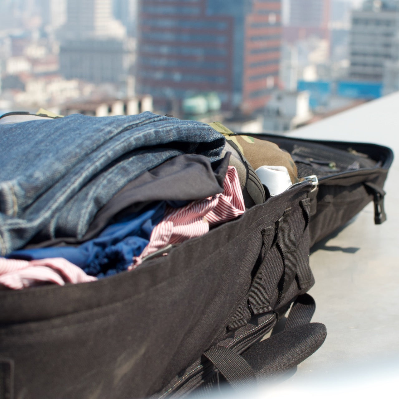 Open suitcase filled with clothes and a GR2 - Cordura ruck on a rooftop, city skyline blurred in the background.