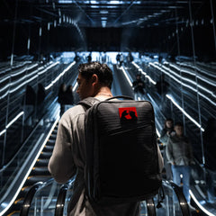 Man on escalator wearing GORUCK rucksack with red patch, modern urban setting, rucking gear