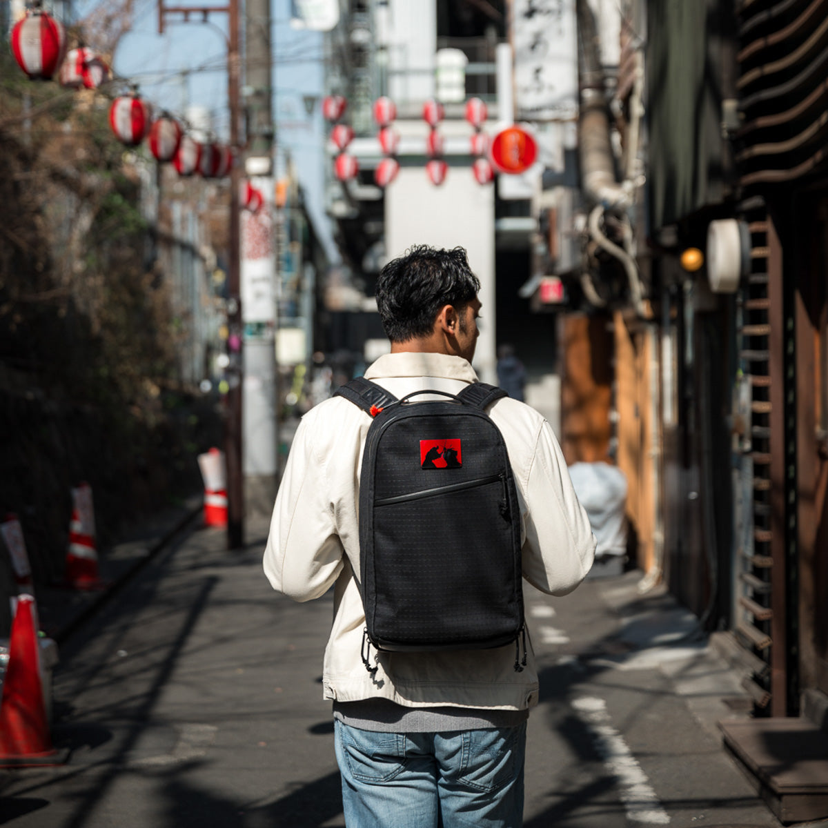 A man with a limited-edition Bullet Ruck x Carryology - GRXC2 Samurai 2.0 backpack walks down a narrow street lined with red lanterns on a sunny day.