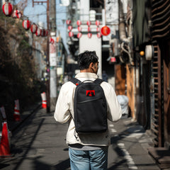 A man with a limited-edition Bullet Ruck x Carryology - GRXC2 Samurai 2.0 backpack walks down a narrow street lined with red lanterns on a sunny day.