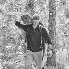 A man carrying a large rock on his shoulder walks through a forested area, wearing Men’s Challenge Shorts - Lightweight ToughDry®, known for being abrasion resistant and fast-drying, along with a cap.
