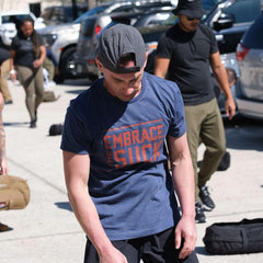 Man wearing navy GORUCK Embrace the Suck tee at outdoor rucking event with others.