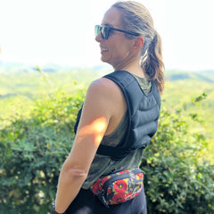 A smiling woman outdoors in a greenery setting wears sunglasses, the Spy Ruck | Women's Weighted Vest, and a colorful waist bag.