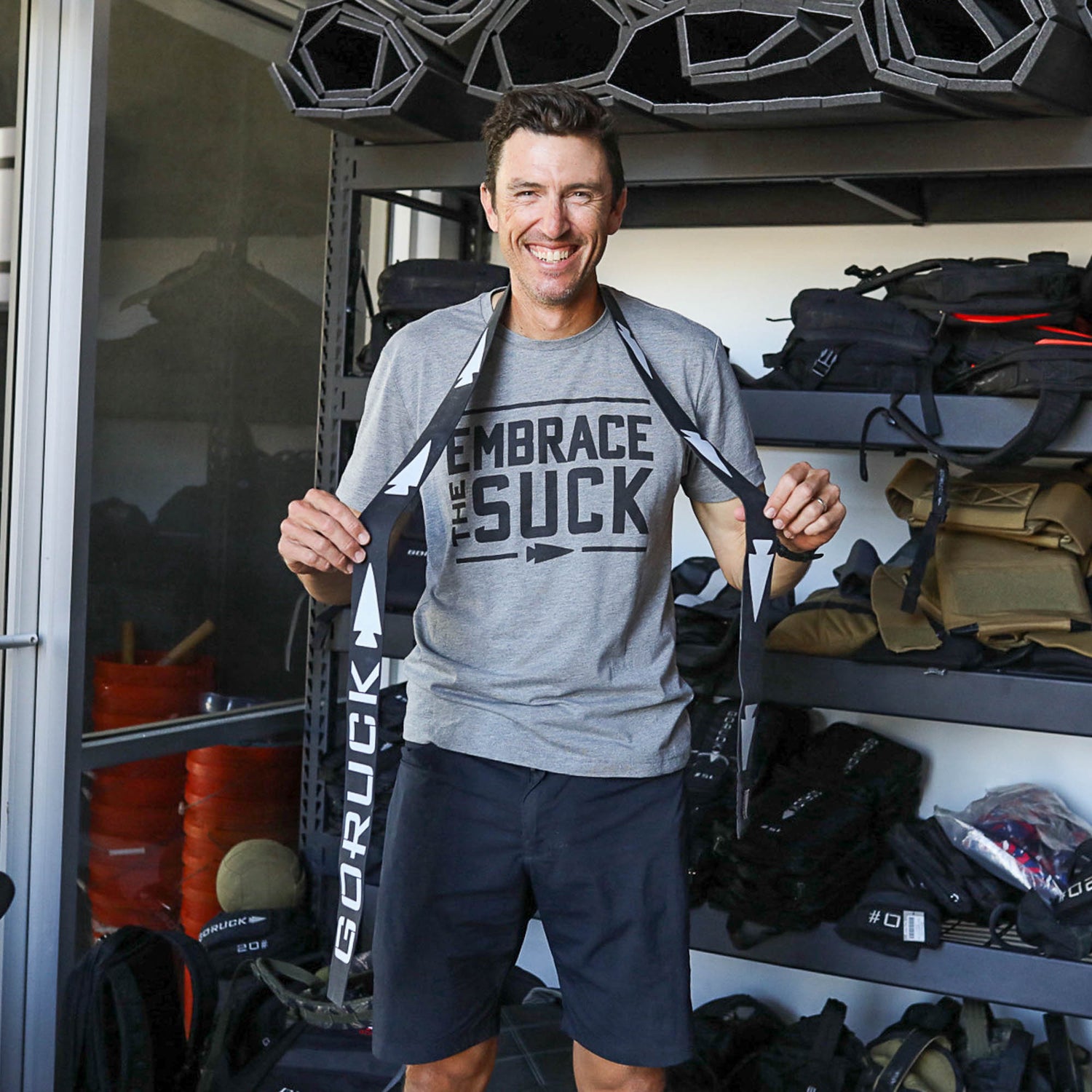 Smiling man in athletic wear holds a GORUCK strap and a Recovery Floss Band, standing before shelves of gear and backpacks to help improve mobility.
