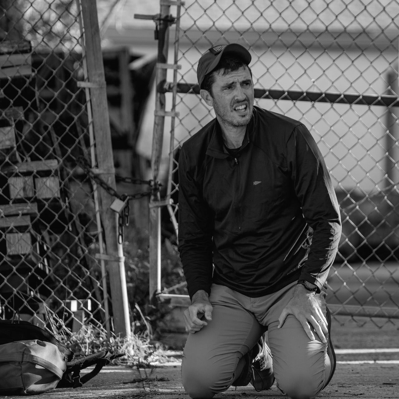 A man kneels by a chain-link fence, looking distressed in black and white—an image that truly embodies the spirit of the Patch - Embrace The Suck.