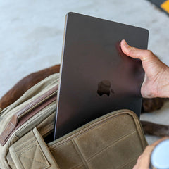 A hand slides a closed MacBook into the laptop compartment of the tan GR2 - Heritage Waxed Canvas backpack, set on a light surface.
