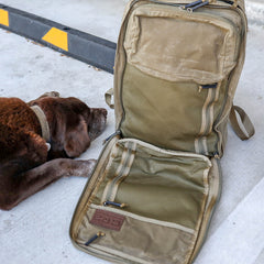 The GR2 - Heritage Waxed Canvas backpack sits open on concrete next to a brown dog by a curb, showcasing its durable laptop compartment that’s perfect for travel.