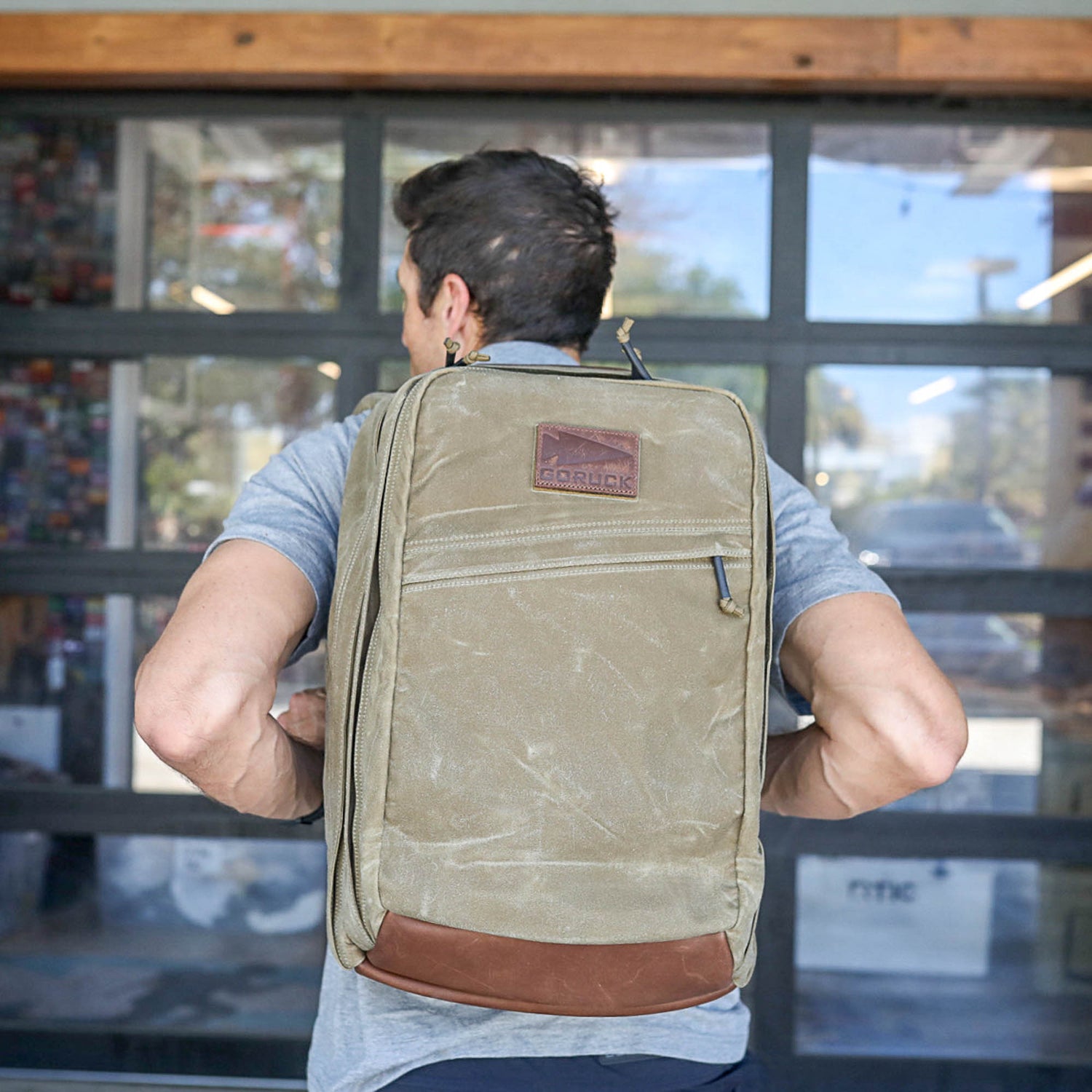 A man stands in front of a glass garage door wearing the GR2 - Heritage Waxed Canvas backpack, featuring green waxed canvas with brown leather accents and a Martexin wax finish.