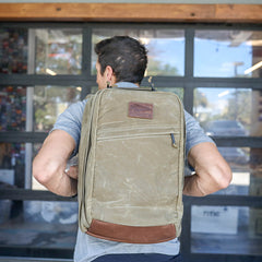 A man stands in front of a glass garage door wearing the GR2 - Heritage Waxed Canvas backpack, featuring green waxed canvas with brown leather accents and a Martexin wax finish.