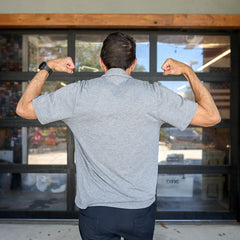Man wearing a gray Men's Polo with Pocket - Tri-BlendX flexes his arms and shows his back while standing in front of glass doors.