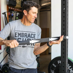 Man wearing a gray "Embrace the Suck" shirt uses a black Recovery Floss Band for muscle compression during his workout in the gym.