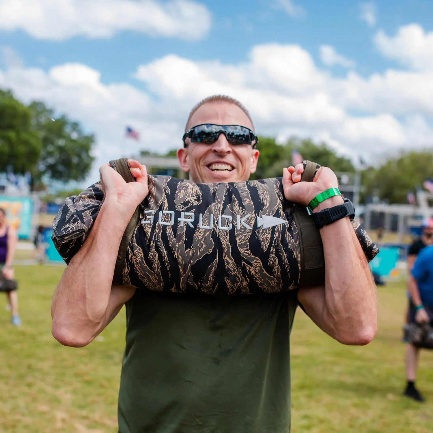 Smiling man outdoors lifting a GORUCK camo sandbag, showcasing tough rucking gear.