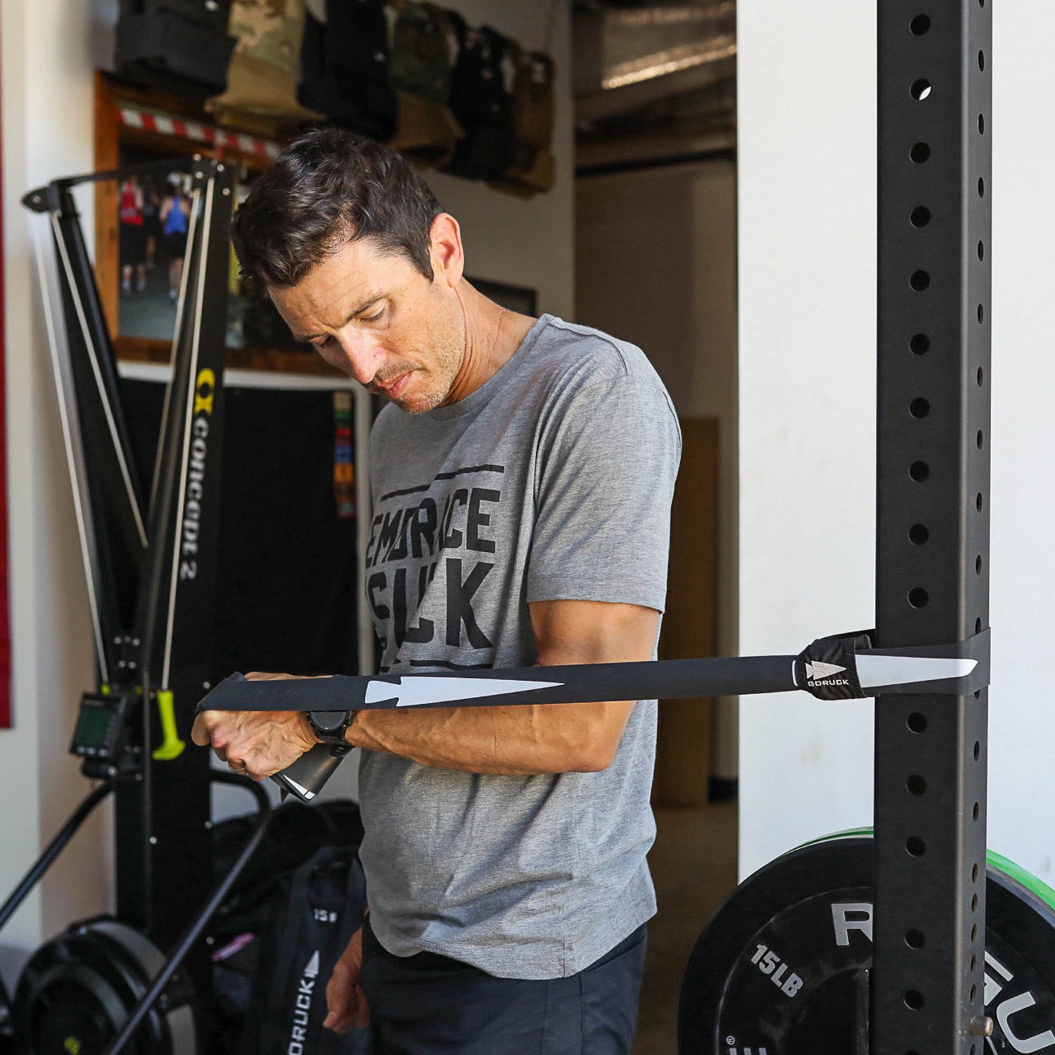 A man in a gym uses the Recovery Floss Band, attached to a squat rack, to perform an arm mobility exercise.
