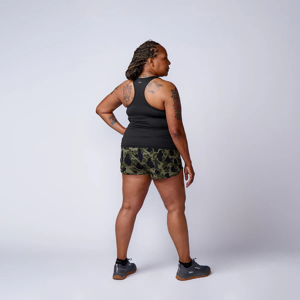 Woman wearing GORUCK camo shorts and black tank top stands in studio, viewed from behind.