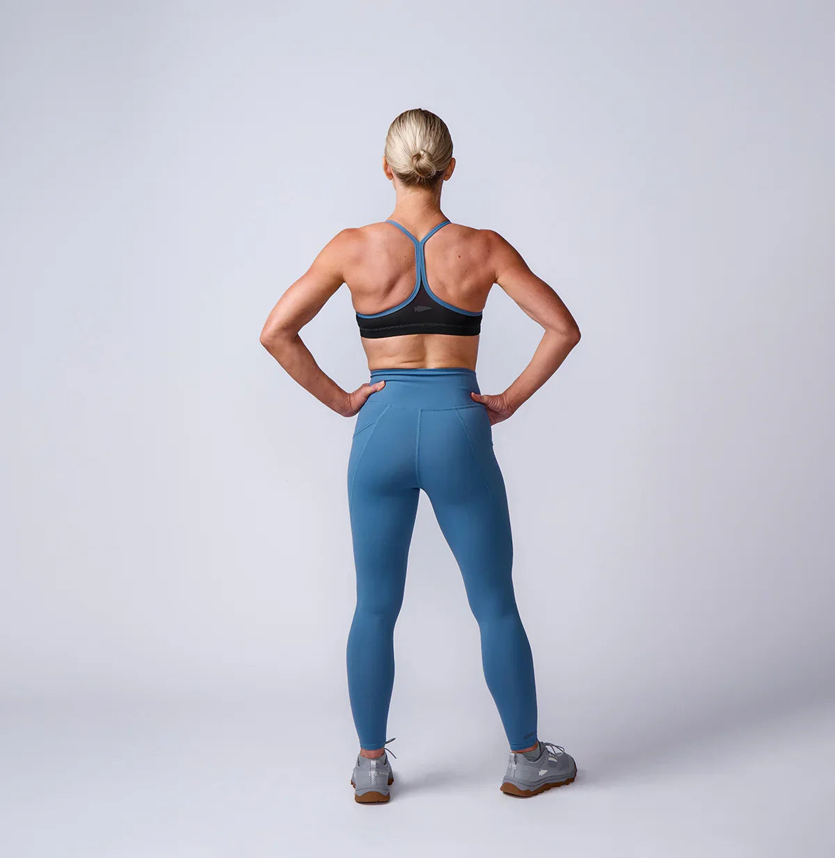 Woman wearing GORUCK blue athletic leggings and sports bra, standing in studio, back view