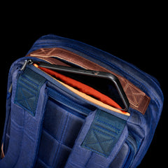 A tablet partially tucked into the top zippered pocket of the GR1 USA - Heritage Waxed Canvas rucksack, featuring blue and brown waxed canvas with orange lining.
