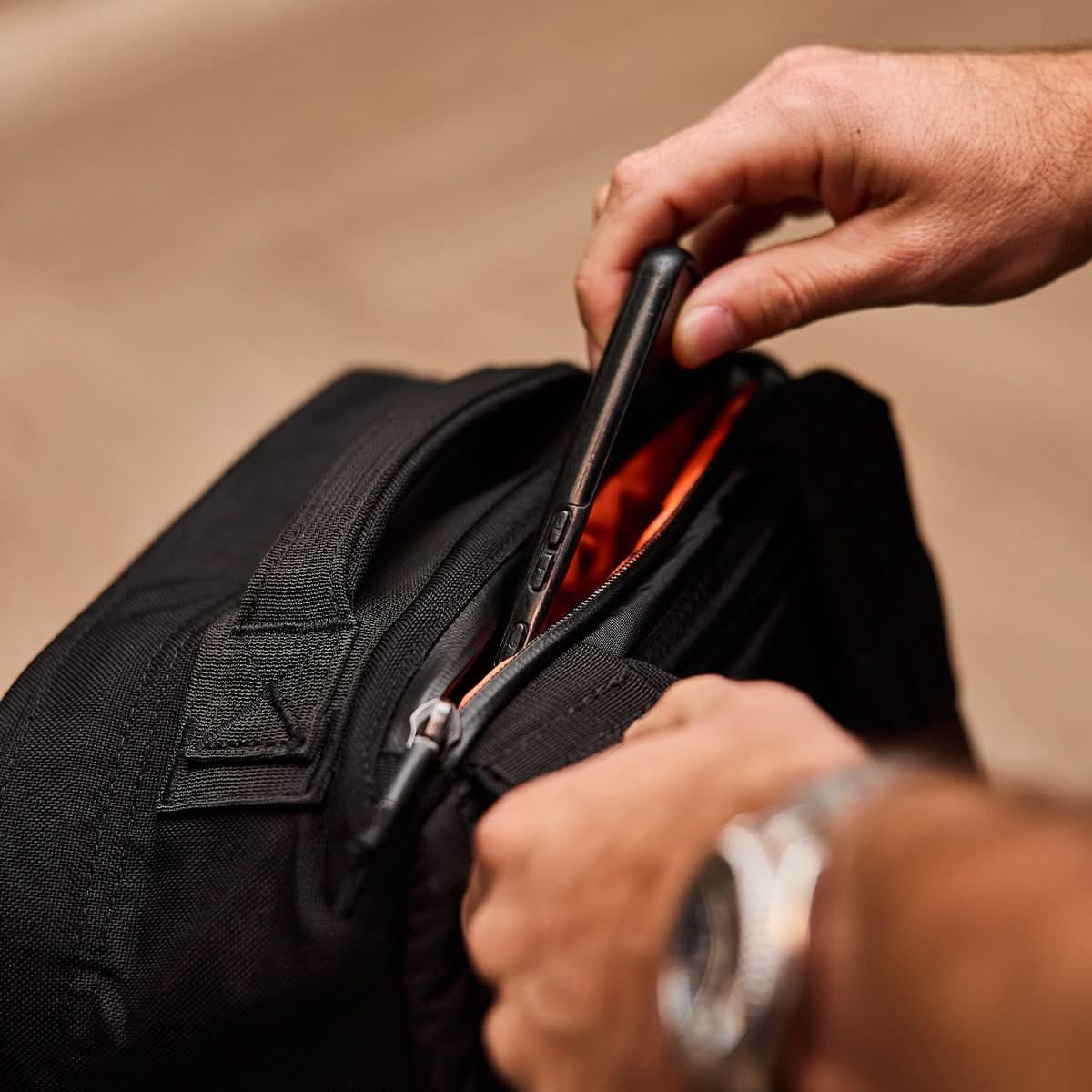 Hand placing phone in black GORUCK rucking backpack with orange interior.