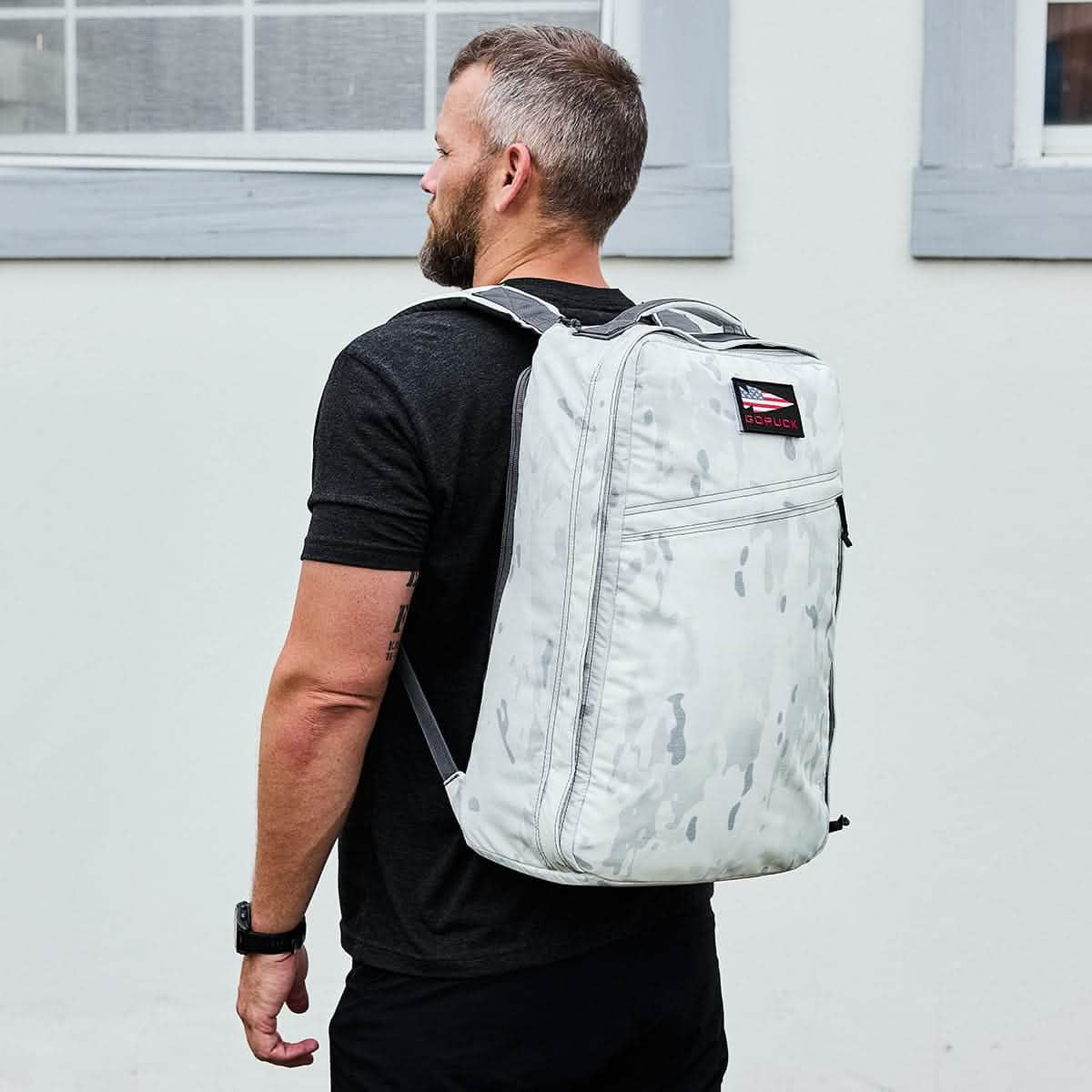 A man in a black shirt wears a light gray GR1 backpack and stands in front of a white building.