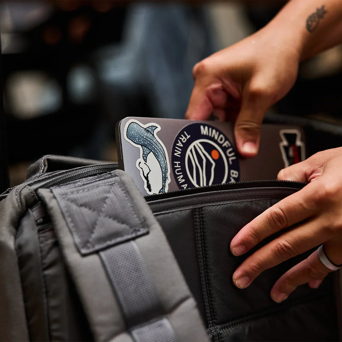 Hands placing a sticker-covered laptop into a GORUCK grey rucksack, close-up view.