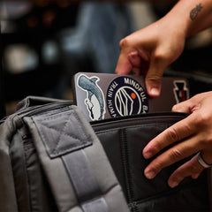 Hands placing a sticker-covered laptop into a GORUCK grey rucksack, close-up view.