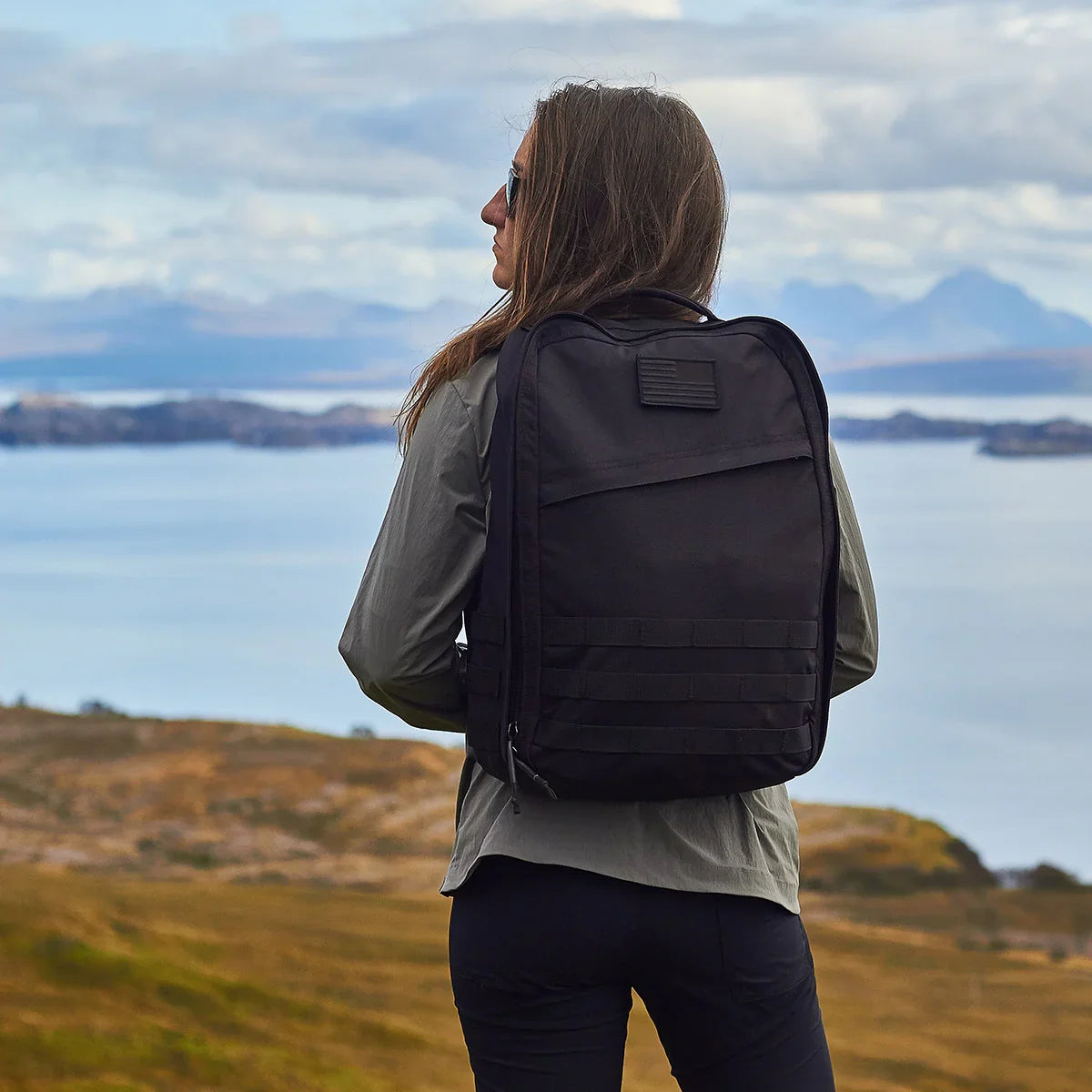 Person hiking outdoors with a black GORUCK rucksack overlooking scenic water and hills