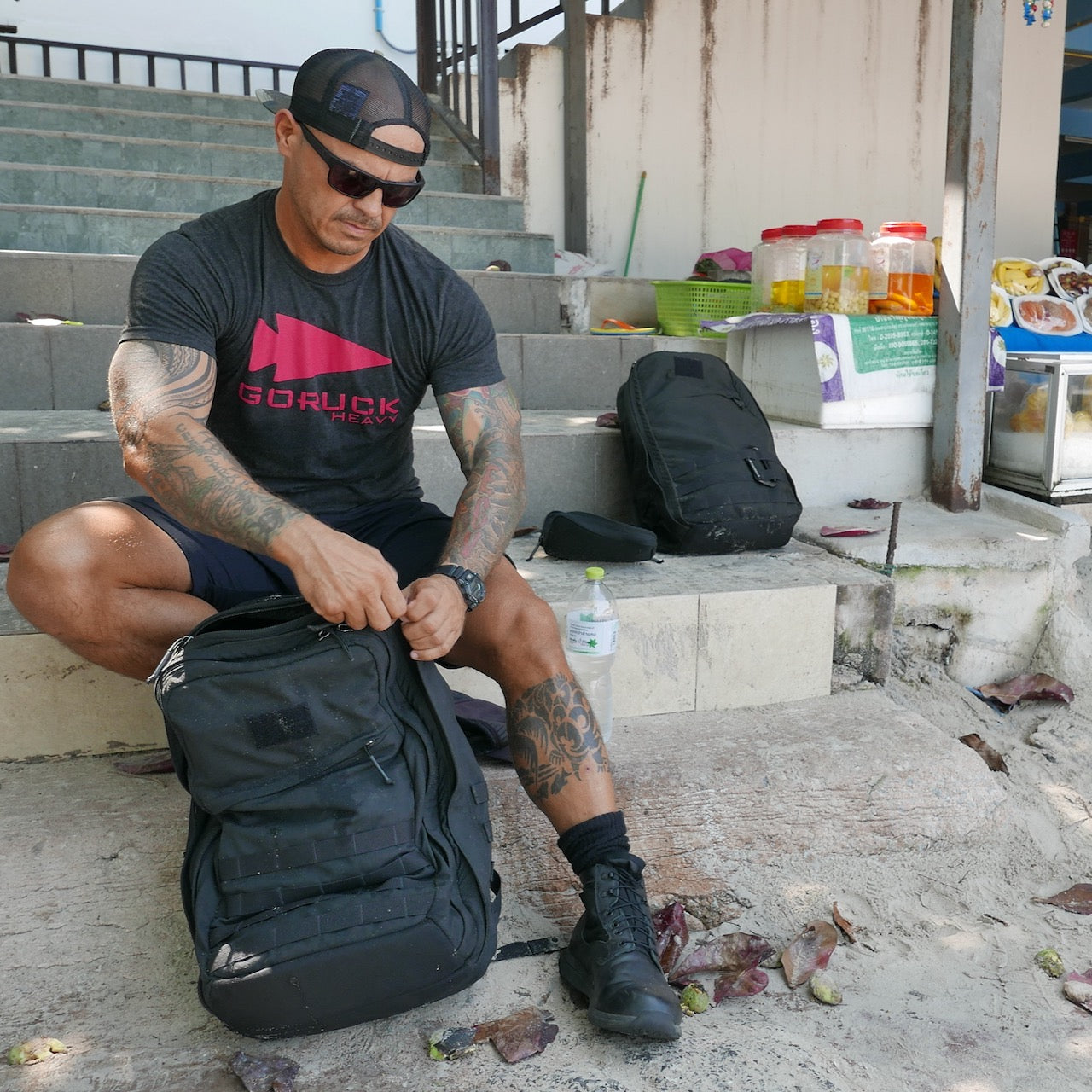 A man in a cap and sunglasses sits on steps, unzipping a black GR2 - Cordura backpack, with an additional carry-on travel bag beside him.