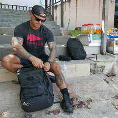 A man in a cap and sunglasses sits on steps, unzipping a black GR2 - Cordura backpack, with an additional carry-on travel bag beside him.