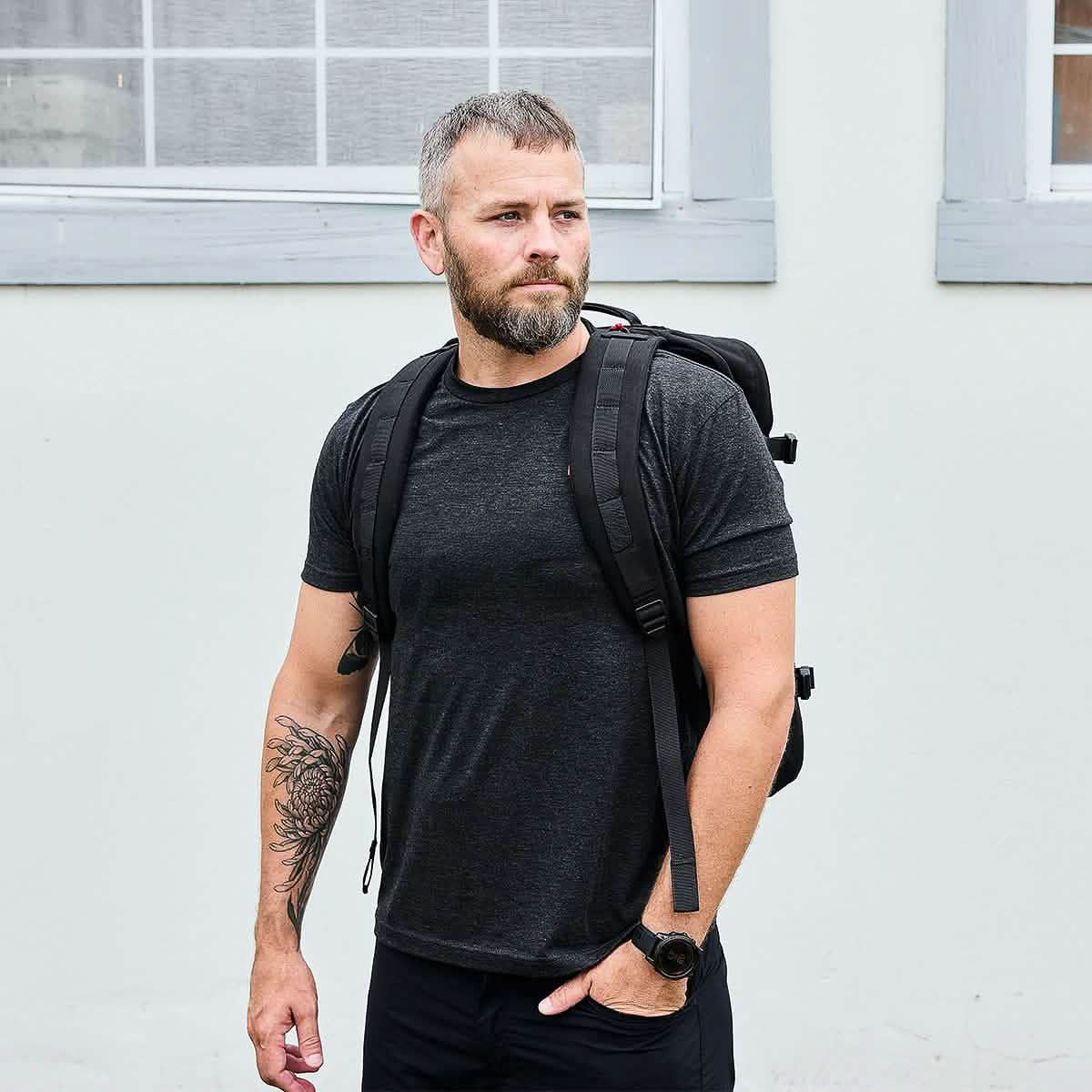 A bearded man in a black t-shirt stands outdoors near a white wall with windows, wearing the GR3 45L travel backpack.