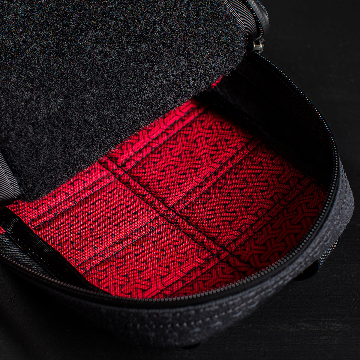 The GRenade x Carryology - GRXC2 Samurai 2.0 features a black exterior with a red patterned interior and zipper, crafted with durable Samurai denim, displayed on a dark surface.