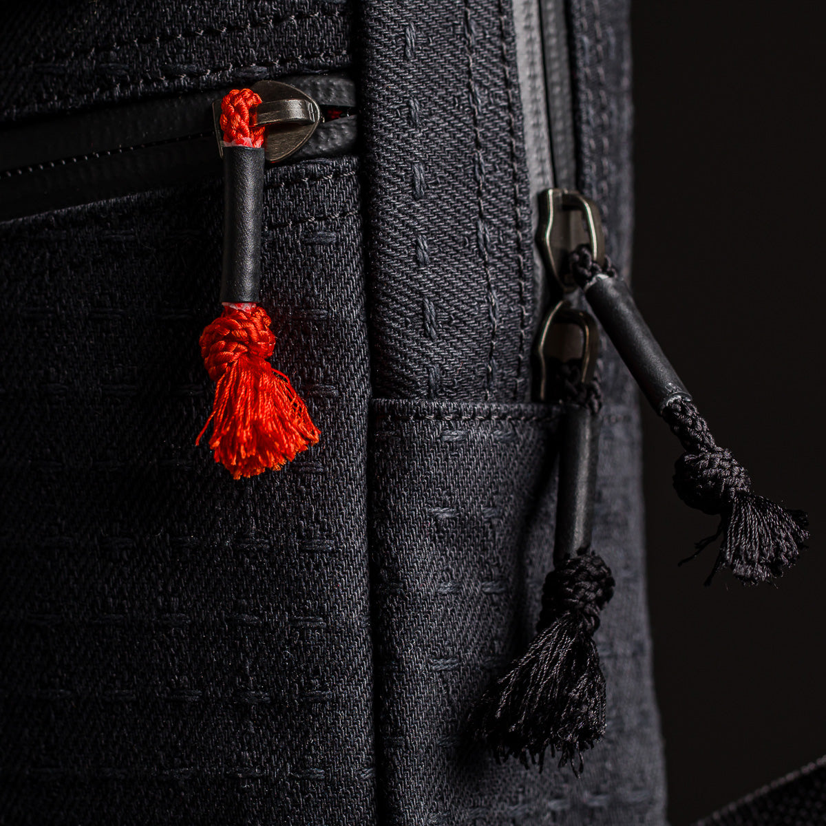 Close-up of the GRenade x Carryology - GRXC2 Samurai 2.0, a black textured tactical sling with red and black tassel zipper pulls.