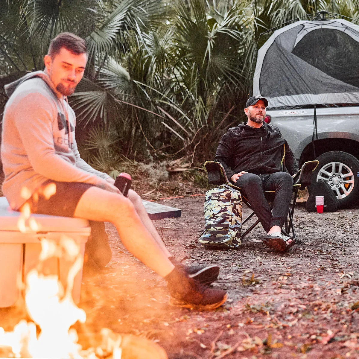 Two men in GORUCK gear sitting by a campfire near a tented vehicle and camo backpack
