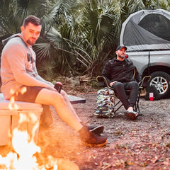 Two men in GORUCK gear sitting by a campfire near a tented vehicle and camo backpack