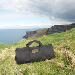 The Gym Bag - Cordura, crafted from durable 1000D CORDURA, rests on grass by seaside cliffs under a cloudy sky—your ideal gym gear organizer.