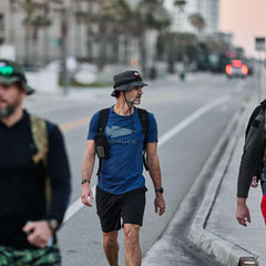 Man wearing charcoal GORUCK boonie hat and blue GORUCK shirt walking outdoors on city street
