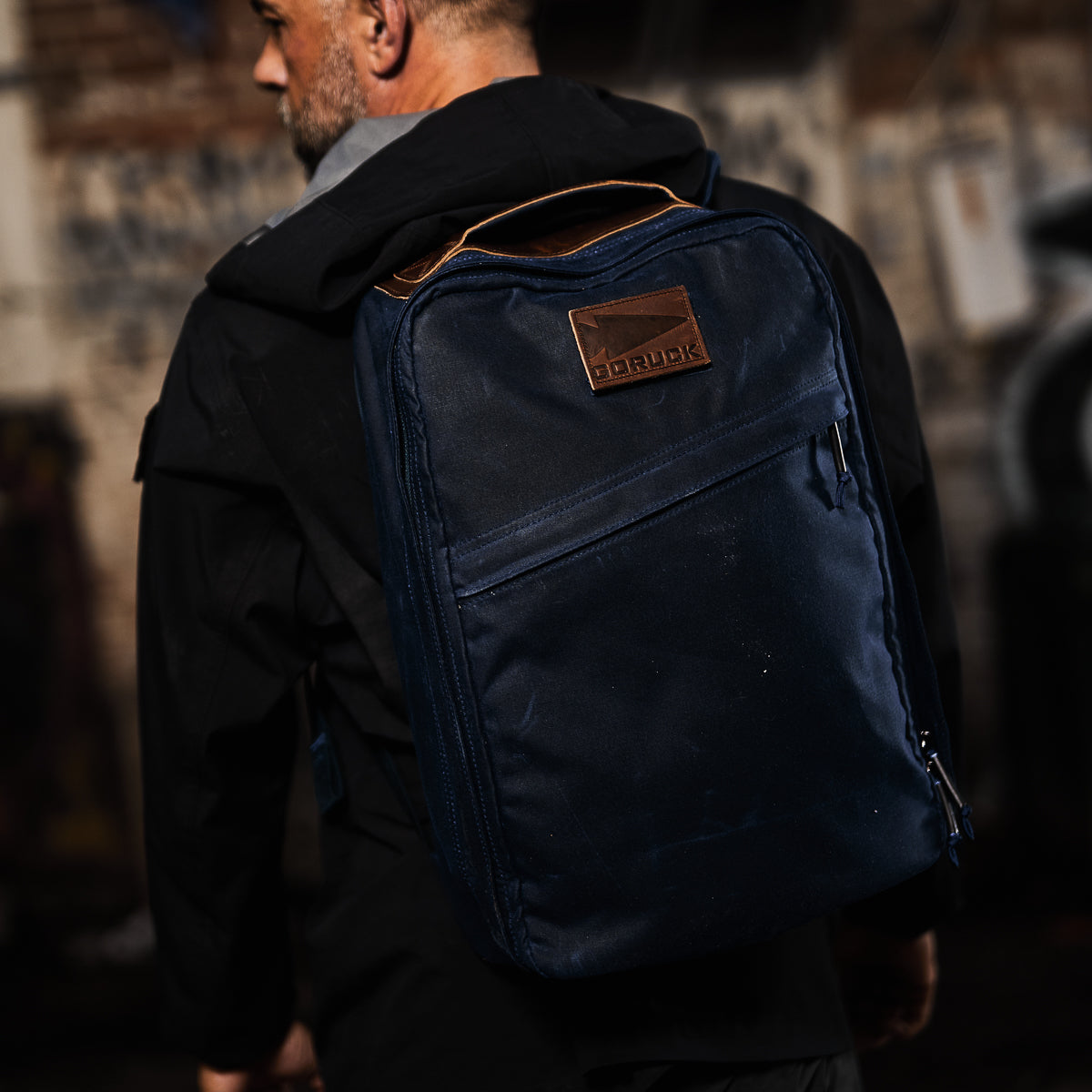 Man wearing a black jacket and carrying the GR1 USA - Heritage Waxed Canvas backpack in navy blue, featuring a brown leather patch on the back.